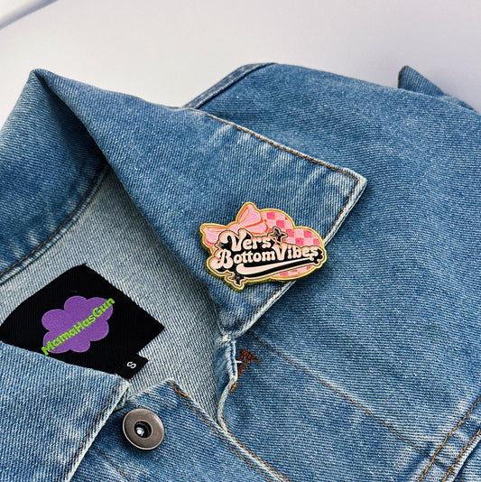 Vers Bottom Vibes Enamel Pin with a bg bow tie on the right top and disco pattern heart on the background. vers bottom vibes written in the middle.
