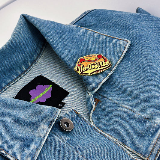 An Enamel pin with a hot dog on the background and vers top written in the middle